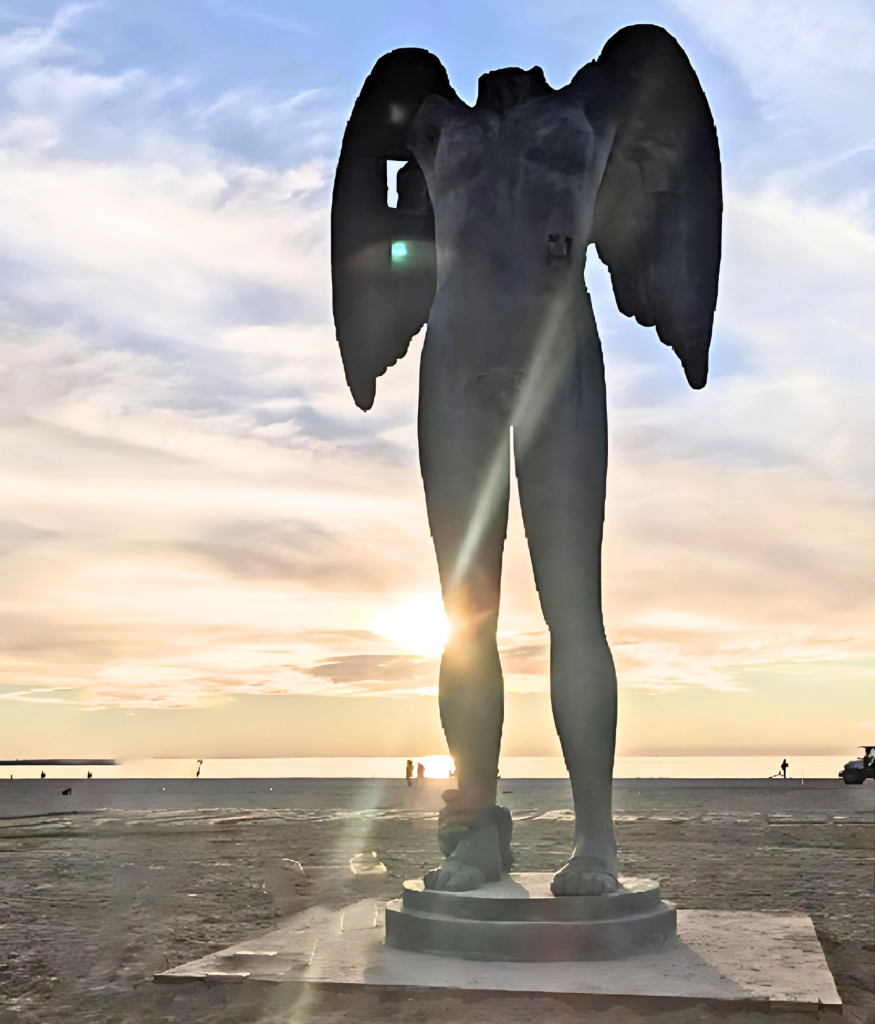 Ikaria statue on a beach with the sun peaking over the left side of the leg and people walking in the background.