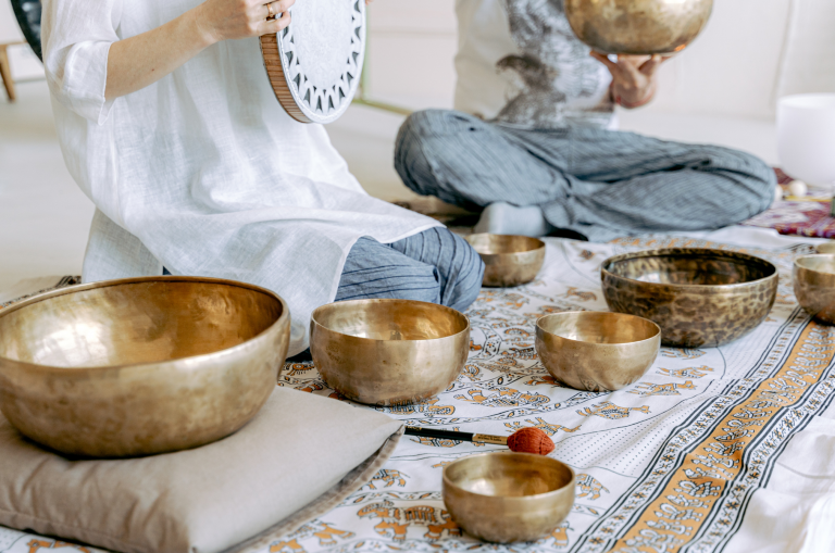 Two people leading a sound bath, one holding a drum and the other a large Tibetan singing bowl and several Tibetan singing bowls in front of them laying over the top a beautiful white tapestry with yellow accents.
