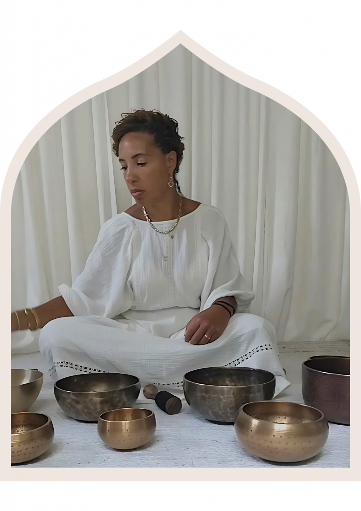 Photo of Lisa Leading a Sound bath. She is wearing a loose white dress and silver jewellery and playing Tibetan singing bowls. The photo is in Islamic Arch frame.